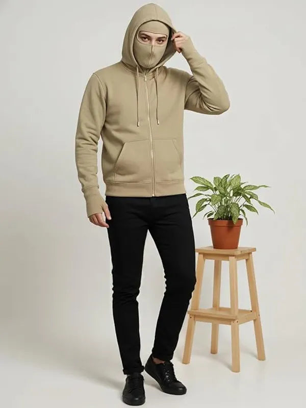 Person wearing a beige hoodie with a face mask, standing next to a wooden stool with a plant on it.