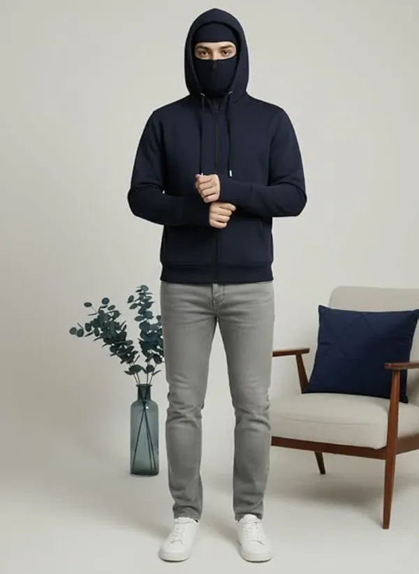 Person wearing a dark hoodie with a face covering in a minimalistic room.