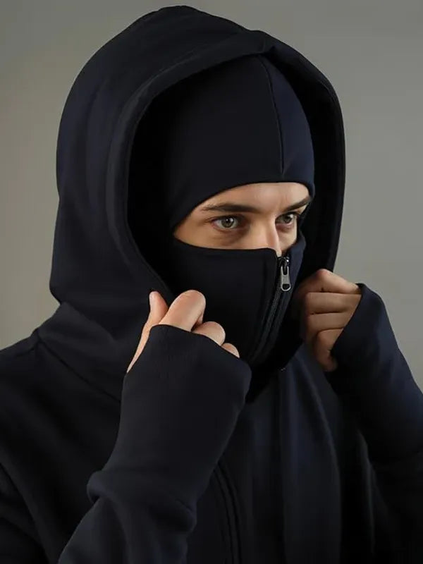 Person wearing a black hooded outfit with a face covering against a gray background