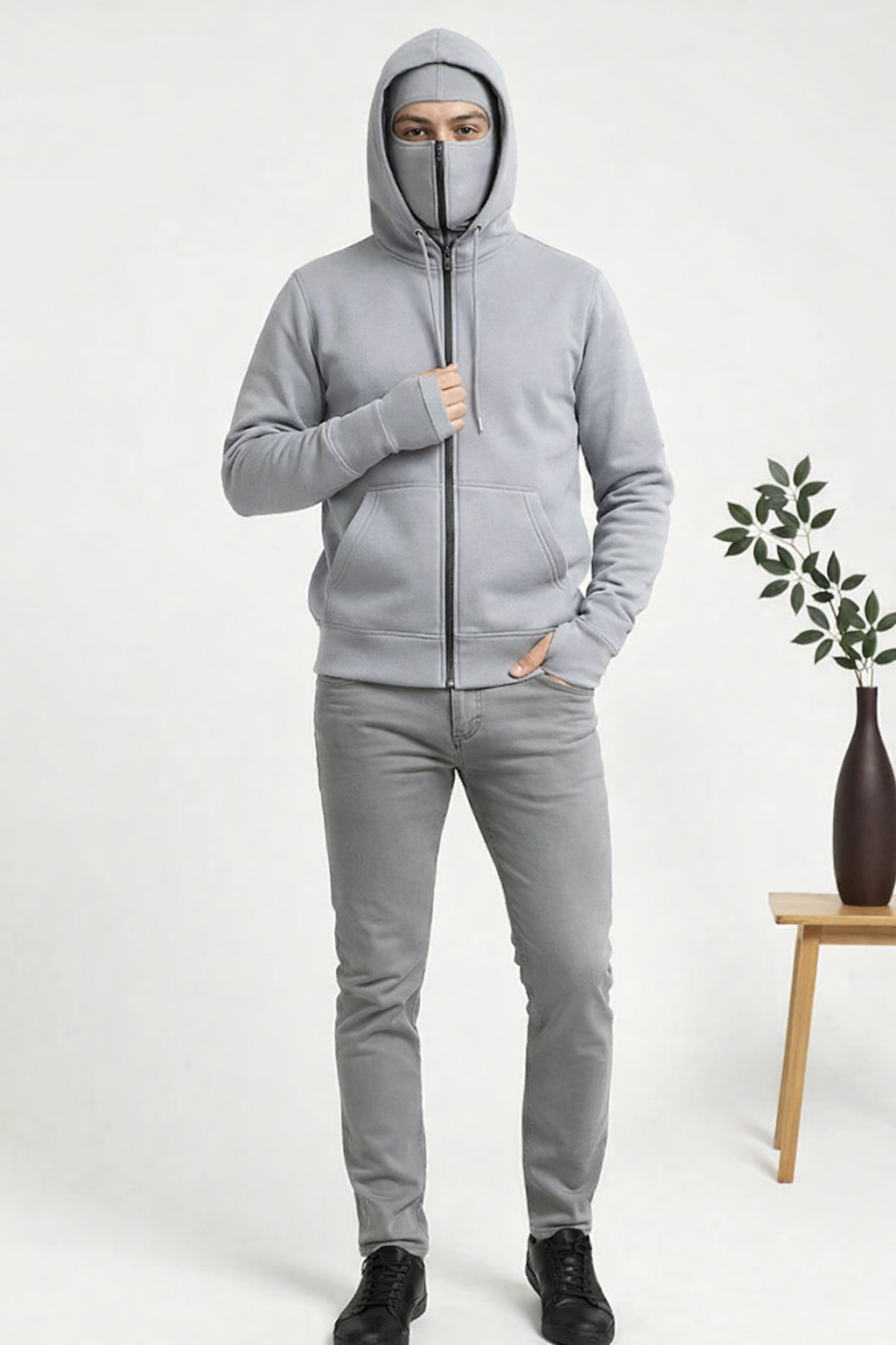 Person wearing a gray hoodie and pants on a white background with a plant and vase.