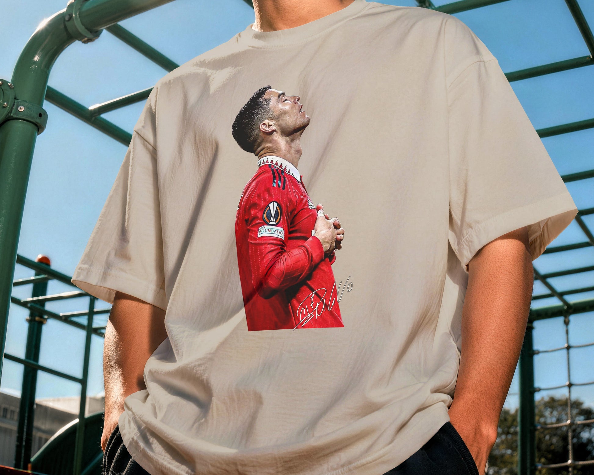 Person wearing a beige t-shirt with a graphic of a person in a red outfit on a playground.